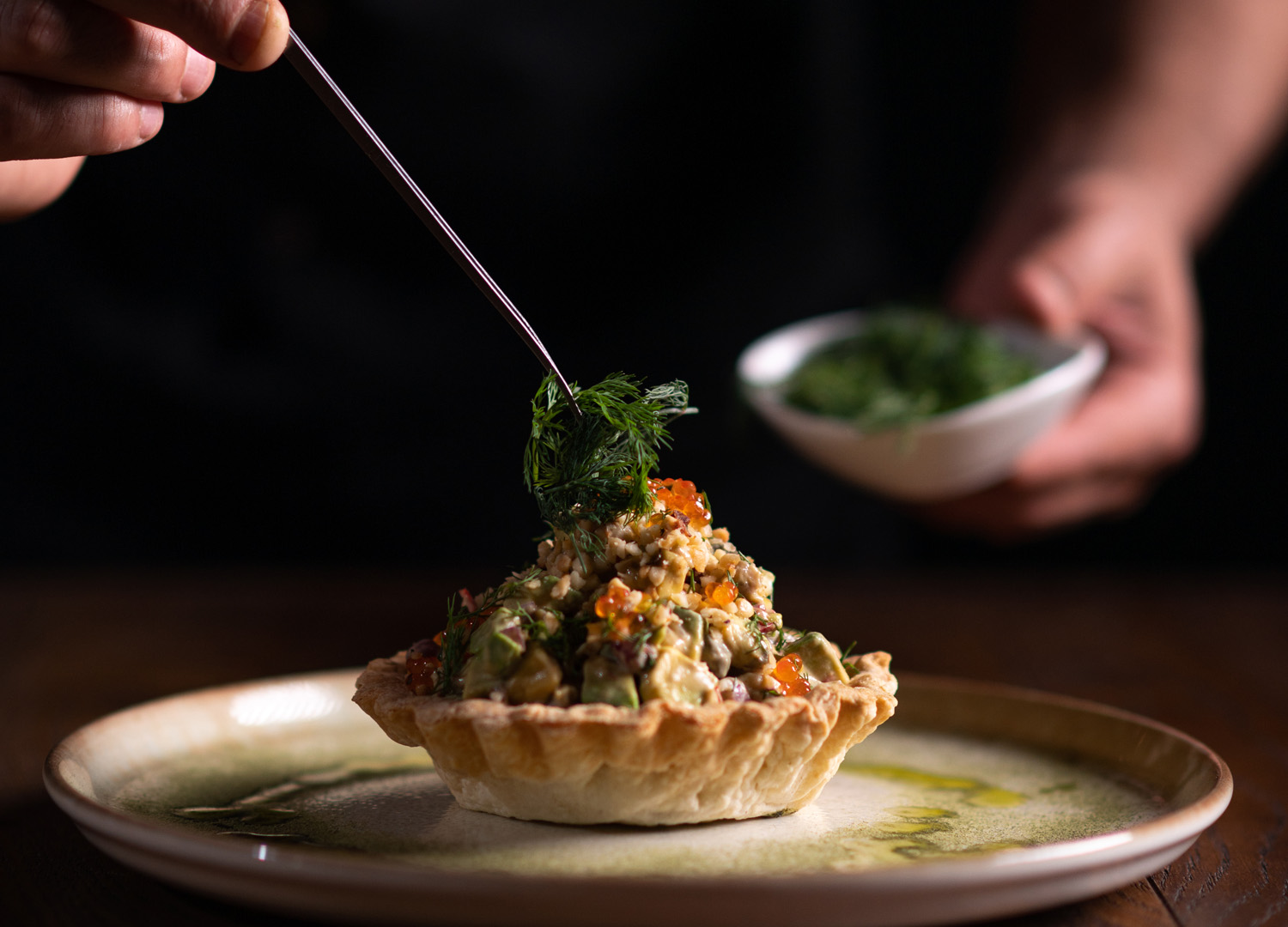 A male chef decorating a plate of tartare di pesce spada with a pinch of dill on a dark background.